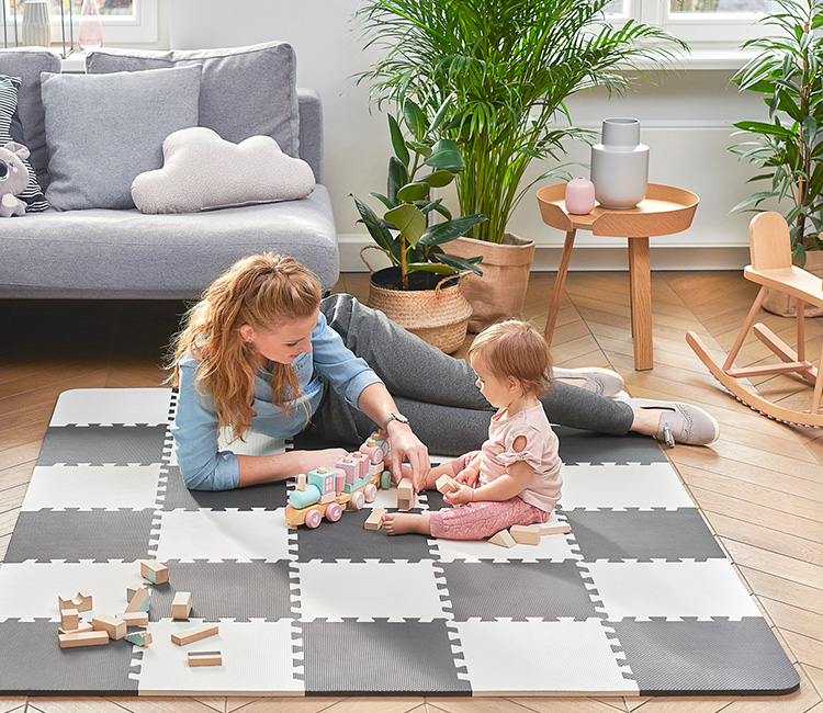Une petite fille d'un an joue avec sa mère sur le tapis en mousse Kinderkraft LUNO dans le salon. Elles construisent un train avec des blocs de bois.