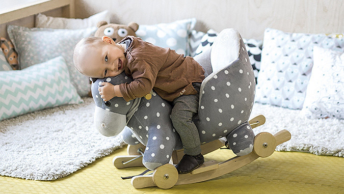 Dans une chambre d'enfant, un petit bébé fait un calin à un cheval à bascule.