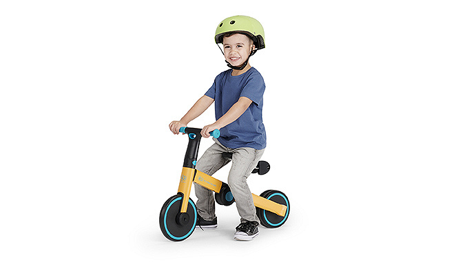 Un garçon de quelques années en t-shirt bleu, jean et chaussures sportives est assis sur un tricycle jaune de la marque Kinderkraft. Il porte un casque.