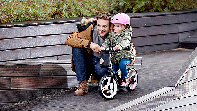 Une fille qui porte un casque rose est assise sur un vélo de la marque Kinderkraft. Son papa la tient et ils regardent tous les deux dans la même directions.