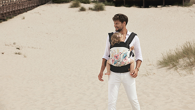 Un papa élégant, habillé dans une chemise blanche est sur une plage. Sur son ventre, dans un porte-bébé de la marque Kinderkraft, une petite fille.