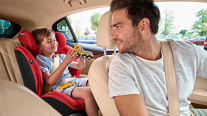 Dans une voiture, un papa-chauffeur se retourne vers son garçon de quelques années assis dans le siège-auto de la marque Kinderkraft, en train de jouer.