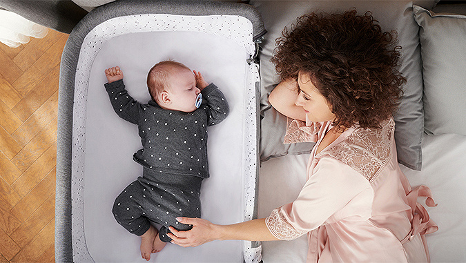 Dans une chambre à coucher, au lit, une maman regarde avec sourire et tient par le pied un petit bébé qui dort dans un lit bébé Kinderkraft qui se trouve à côté.