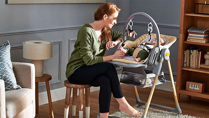 Une maman souriante est assise sur une petite chaise dans une chambre. Elle tient un bébé par le pied, qui dort à côté d'elle dans un siège Kinderkraft. 