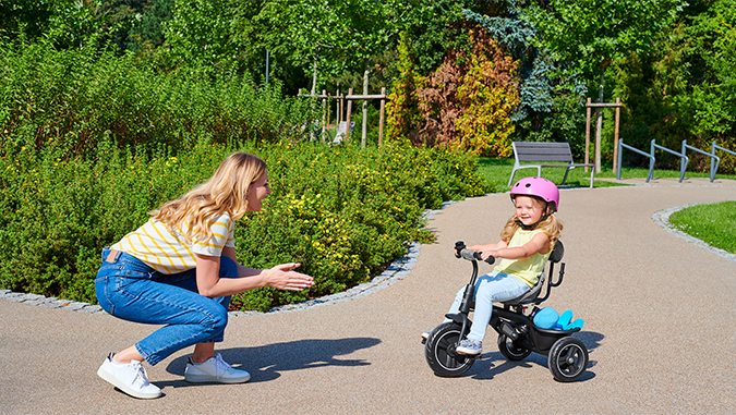 Sur un chemin au parc lors d'une journée ensoleillée, une fille porte un casque rose et roule sur un tricycle de la marque Kinderkraft dans la direction de sa maman qui sourit.
