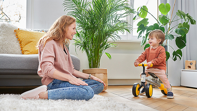 Un petit garçon roule sur un tricycle de la marque Kinderkraft dans sa chambre. Sa maman est assise par terre et le regarde avec sourire.