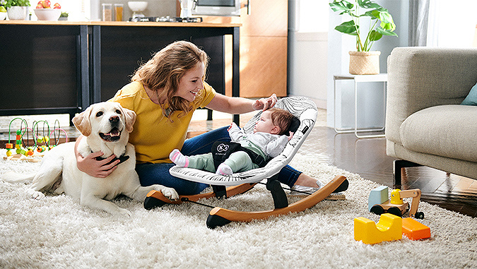 Une maman est à la maison sur un tapis et caresse un petit enfant allongé dans un transat de la marque Kinderkraft. Elle tient un chien avec son autre main.