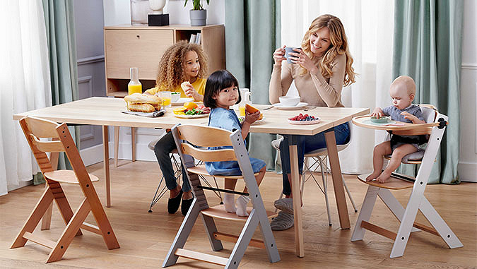 Une maman et trois enfants prennent le petit déjeuner sur une table. Les deux plus petits enfants sont assis sur des chaises spéciales Kinderkraft.