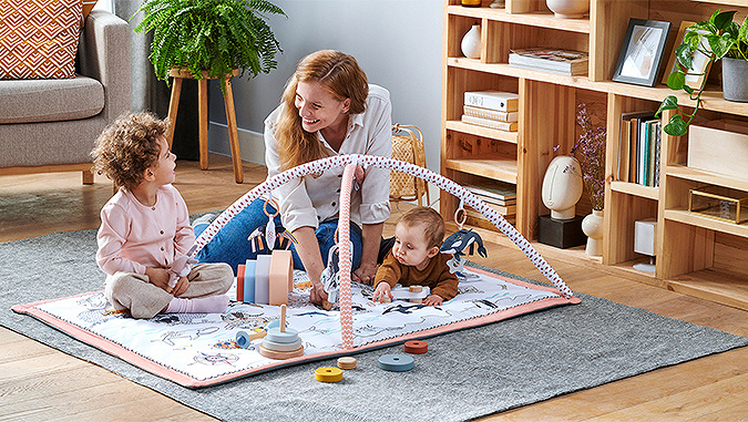 Dans un appartement, sur un tapis, se trouve un tapis éducatif déplié de la marque Kinderkraft. Dessus, une maman, un enfant de quelques années et un nouveau-né jouent ensemble.
