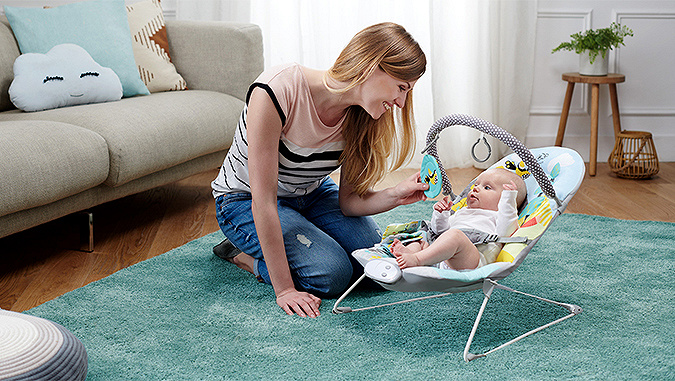 Une maman est agenouillée sur le tapis et sourit, en montrant le jouet au bébé allongé dans un transat Kinderkraft. L'enfant la regarde avec intérêt. 
