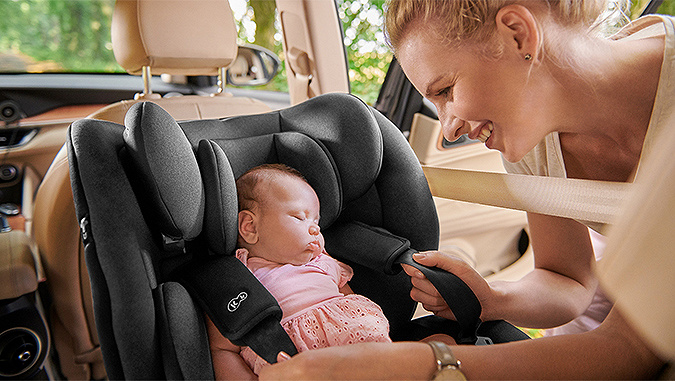 Un nouveau-né dort dans un siège-auto gris de la marque Kinderkraft orienté dos à la route, et sa maman, souriante, lui met les ceintures.