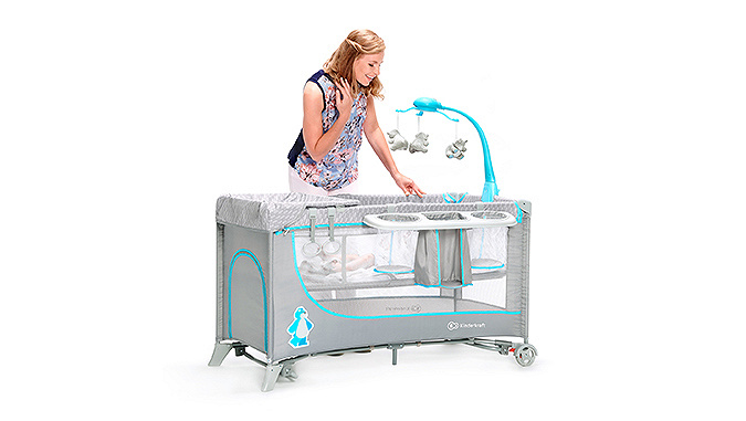 Dans un lit gris de la marque Kinderkraft avec un carroussel suspendu avec jouets, un bébé. Une maman souriante se penche sur lui.