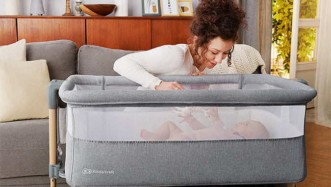Dans un apartement, une maman se penche avec sourire sur un bébé qui joue allongé dans un lit enfant de la marque Kinderkraft.