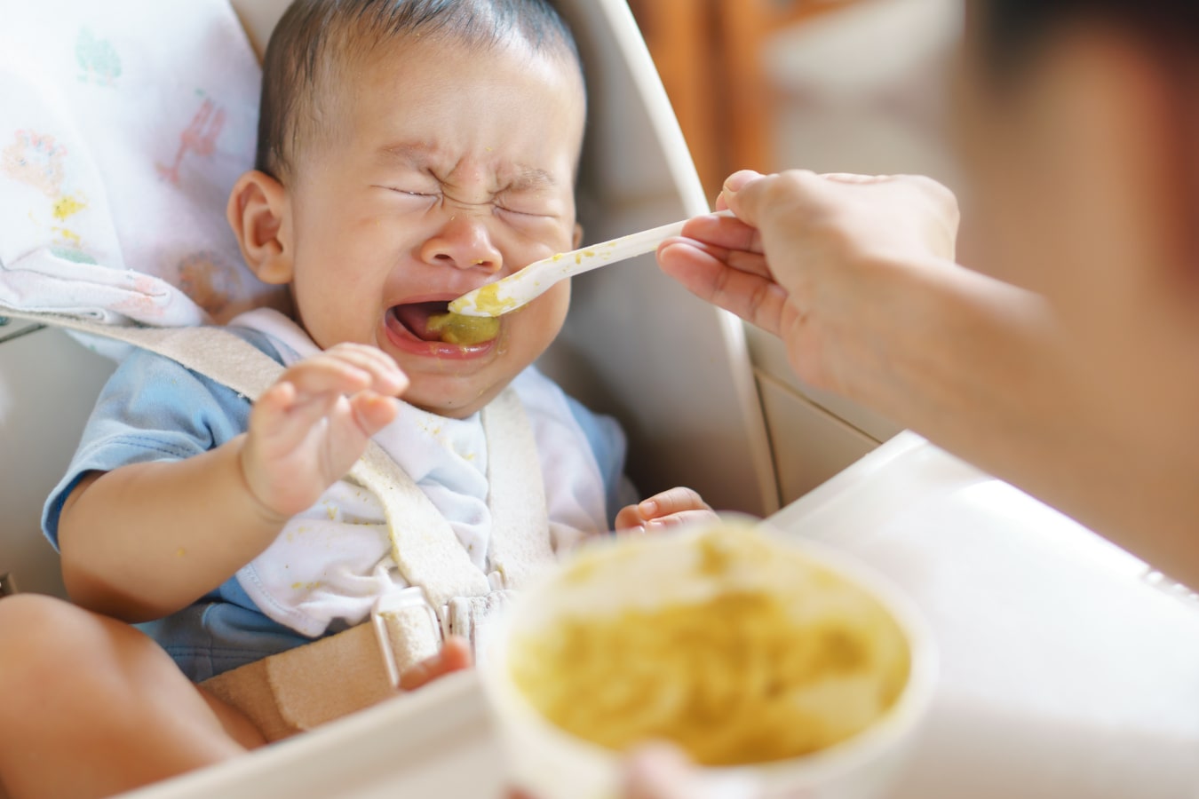 Un bébé est assis dans une chaise haute, il refuse de manger à la cuillère et pleure.