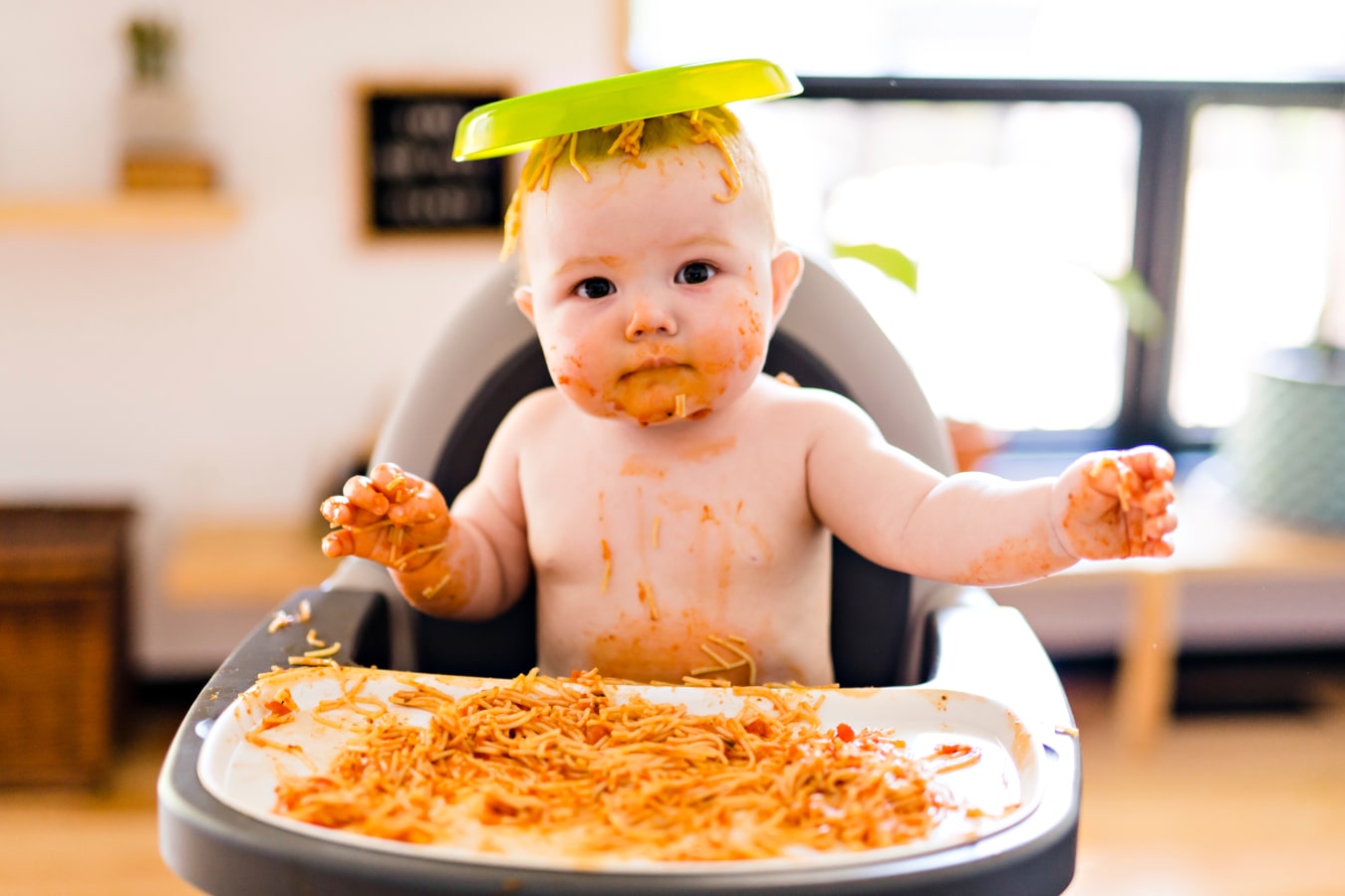Un bébé, sale à cause des spaghettis, est assis sur une chaise haute, avec une assiette de nourriture sur la tête.