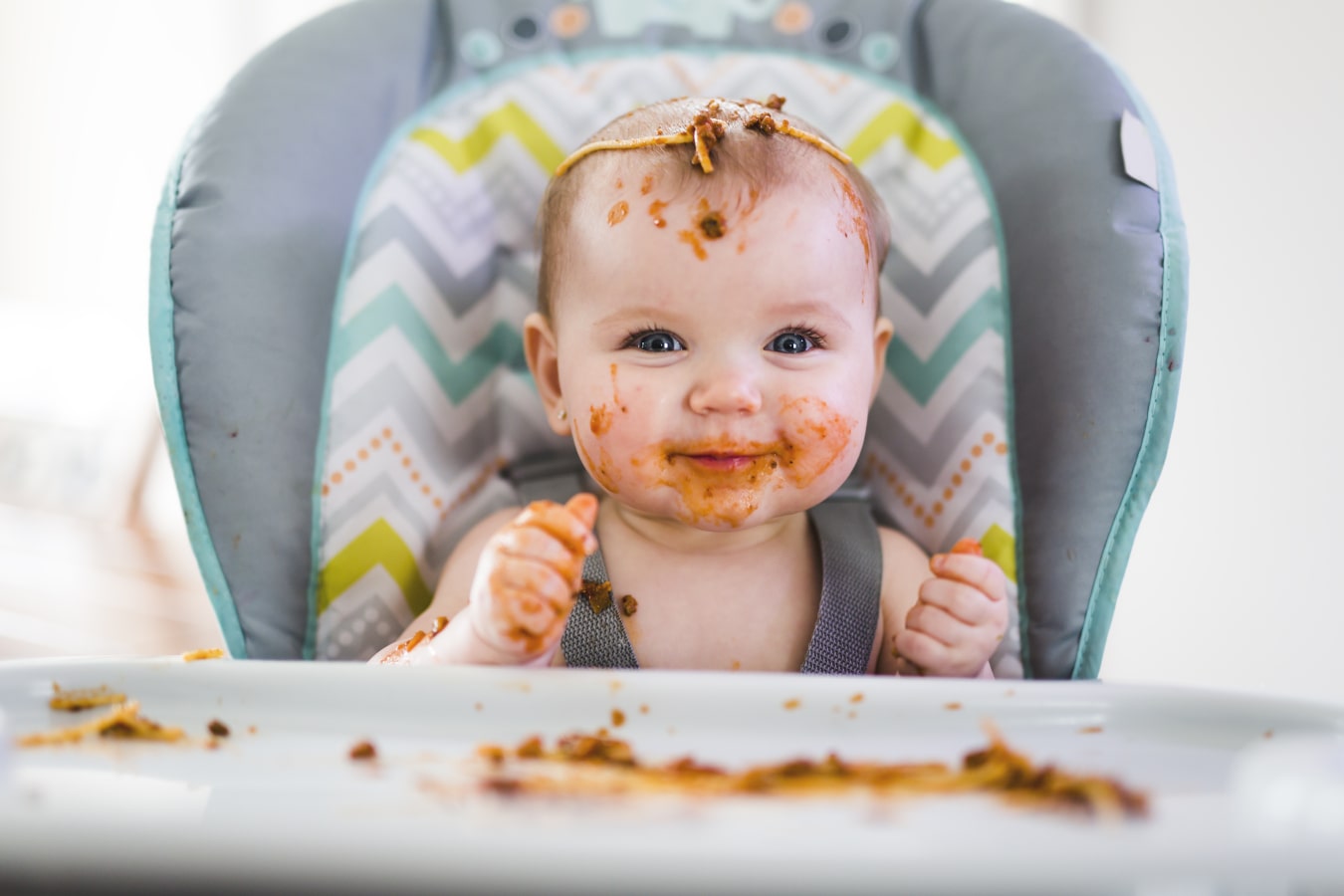 Un bébé, sale à cause des spaghettis, est assis dans une chaise haute. Il est heureux et souriant, il y a beaucoup de nourriture sur lui et sur le plateau.
