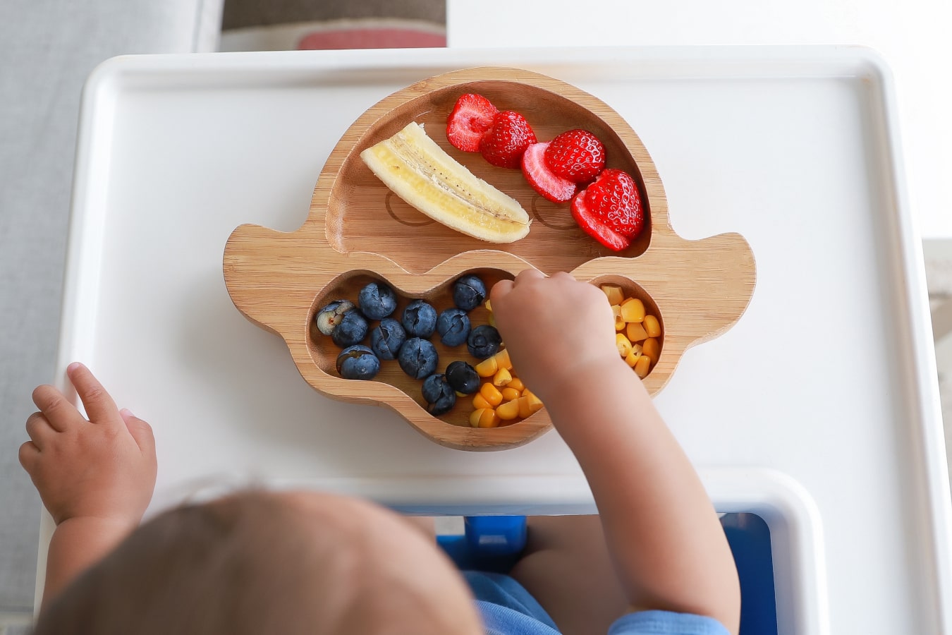 Sur l'assiette en bois à compartiments, on peut voir des fruits et légumes colorés : banane, myrtilles, maïs et fraises. L'enfant attrape la nourriture