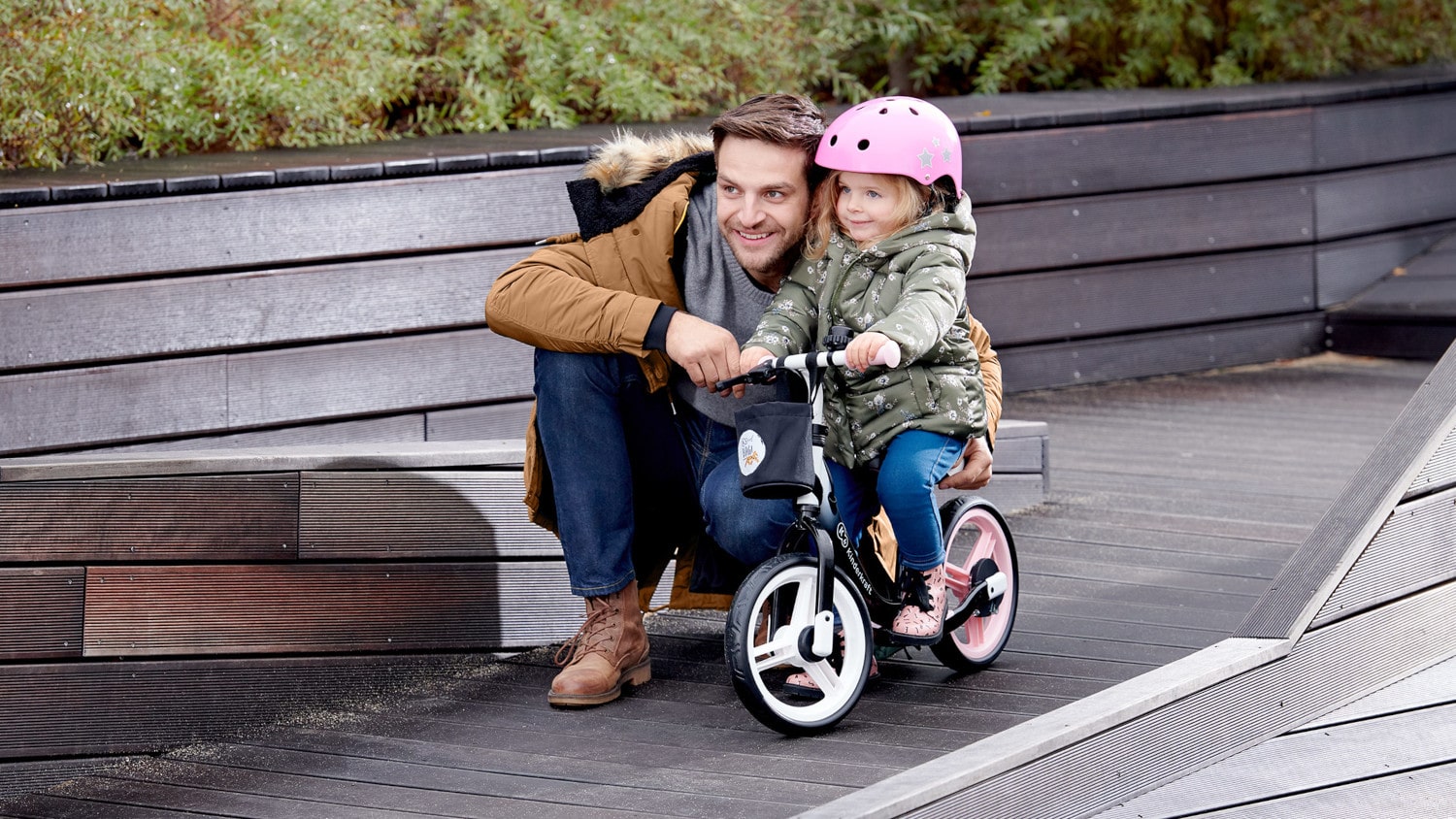 En automne, un père dehors s'accroupit à côté d'une fille assise sur une draisienne noire et rose à deux roues avec un casque sur la tête.