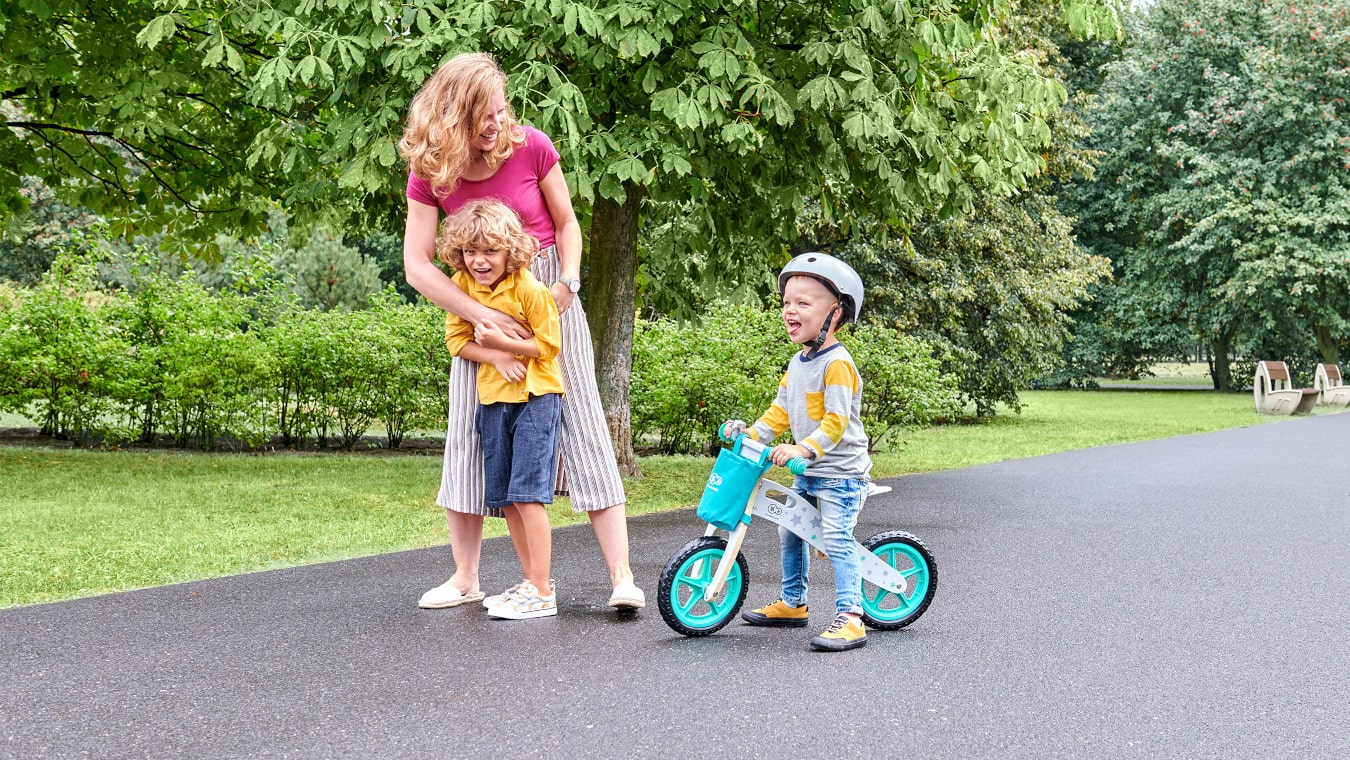Une maman se promène dans le parc avec un enfant sur une draisienne à deux roues turquoise et avec un deuxième enfant plus âgé.