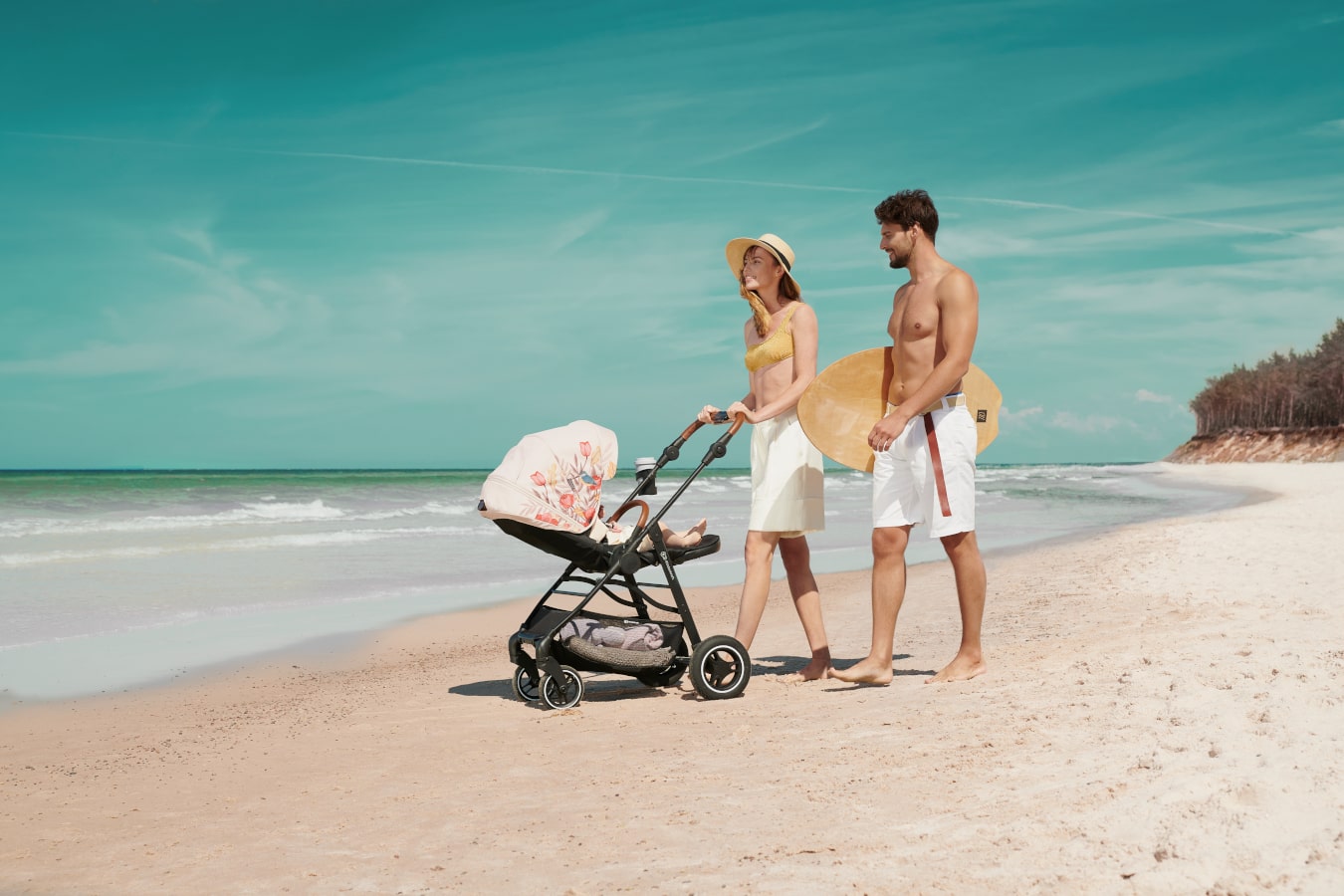 Une maman et un papa se promènent sur la plage avec un bébé allongé dans une poussette noire avec une capote beige.