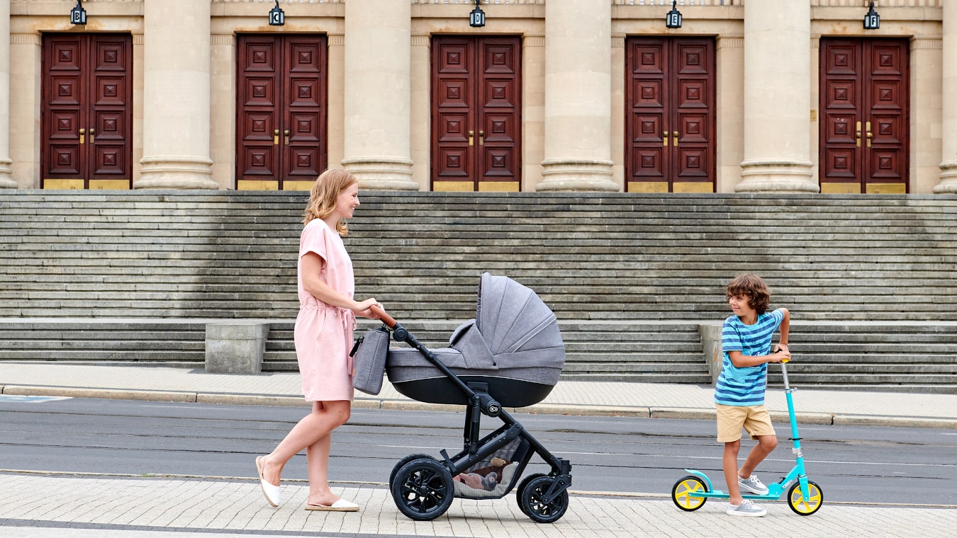 Une maman se promène dans la ville avec une poussette grise et avec un bébé sur une trottinette turquoise à deux roues.