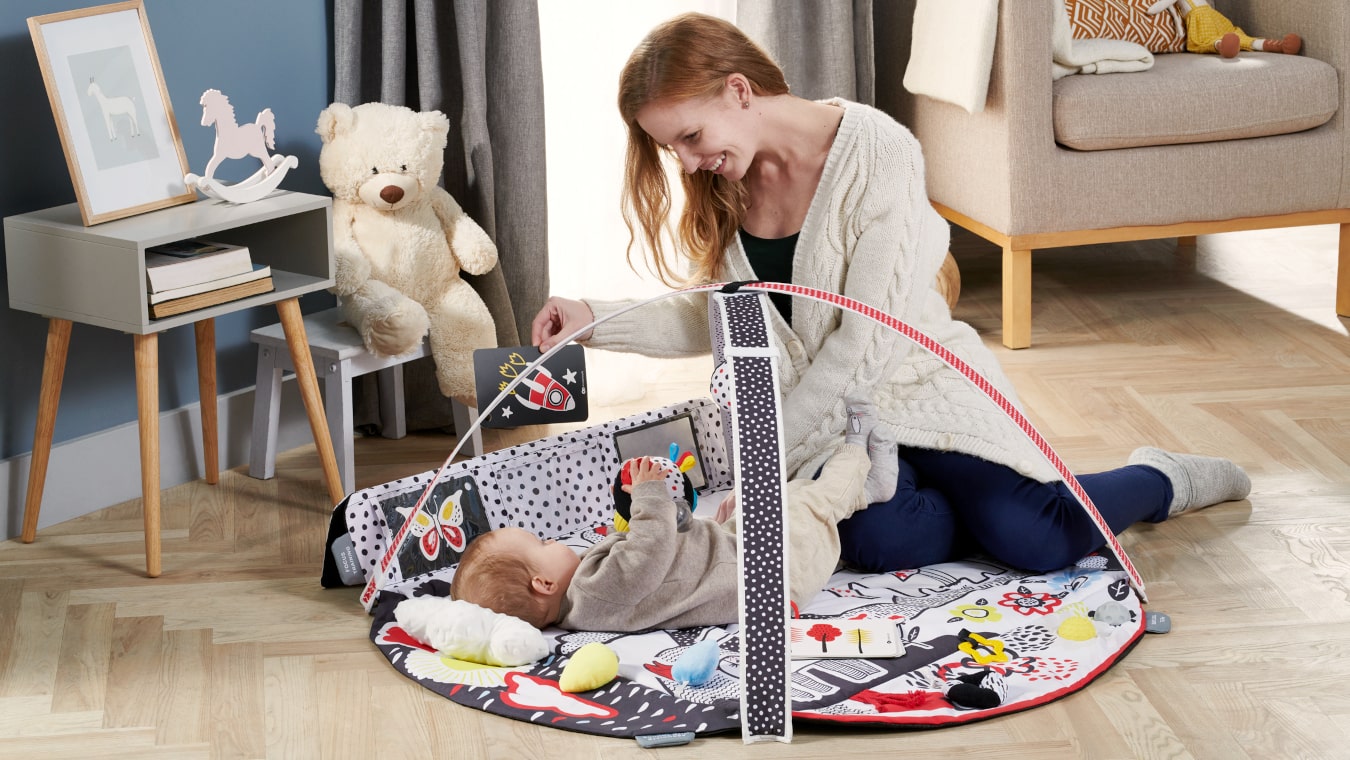 Une mère souriante dans son appartement joue avec un enfant allongé sur un tapis sensoriel coloré, lui montrant une image contrastée.