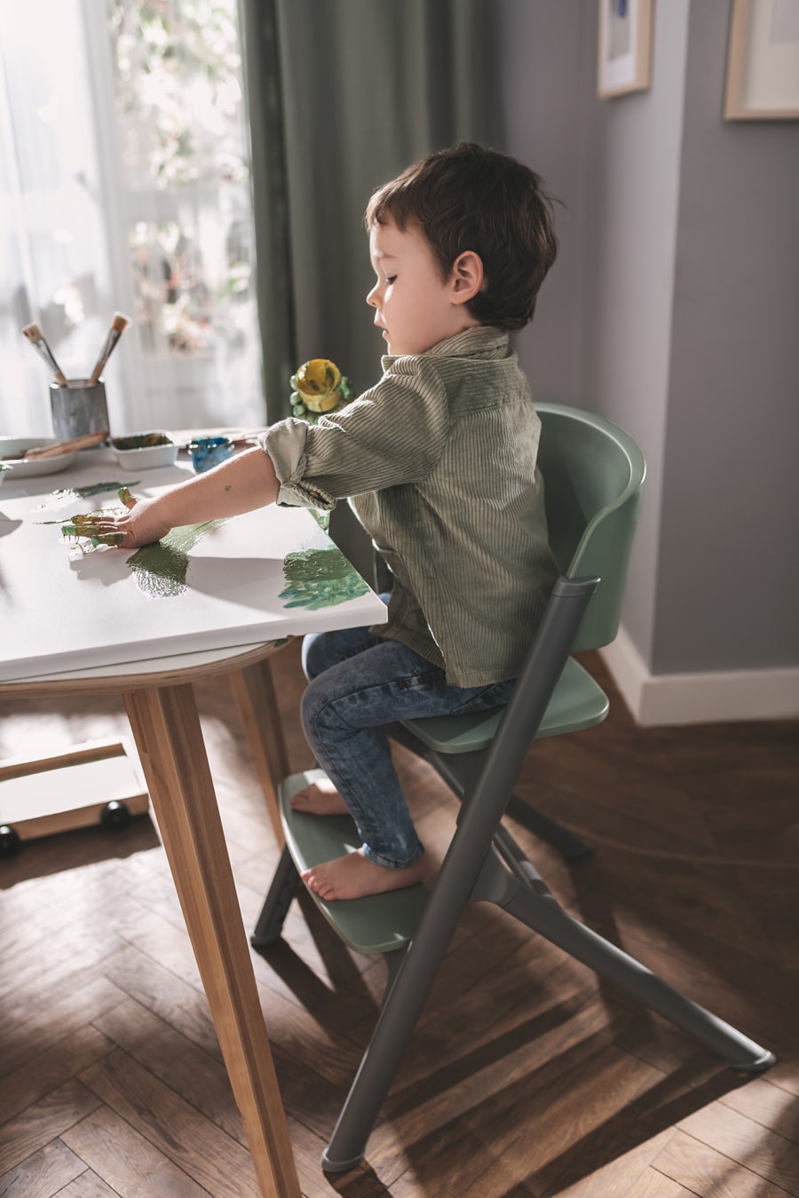 Le petit garçon est assis dans la chaise haute LIVY de Kinderkraft, peignant avec ses doigts sur la toile, il est concentré et détendu