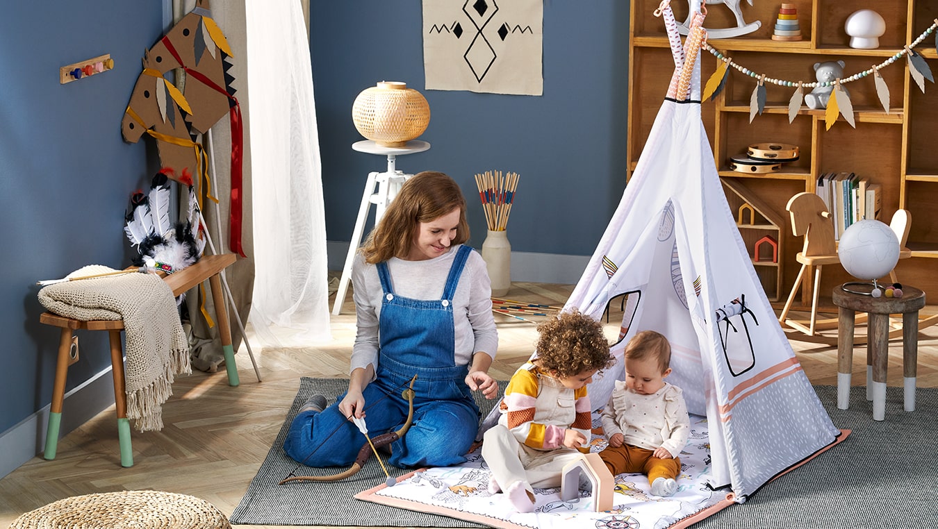 Deux enfant jouent dans la tente tippy kinderkraft, la maman est assise à côté et tient un arc avec des jouets, tous rient et passent un bon moment.