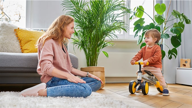 La maman sourit à son enfant qui roule en vélo à trois roues cutie kinderkraft. Ils sont à la maison, en arrière-plan on voit des plantes vertes et un canapé gris. L’enfant sourit.