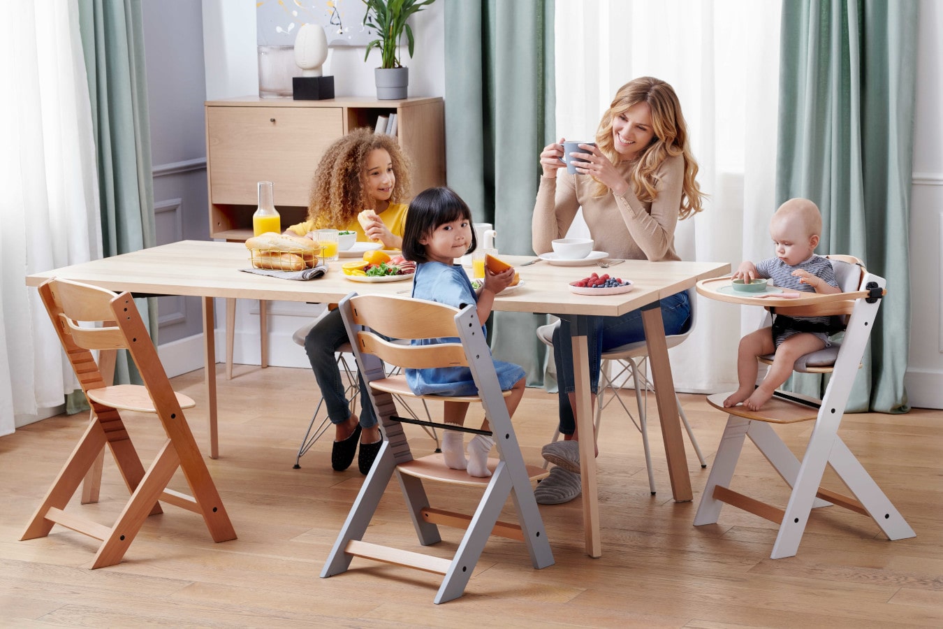 La maman et trois enfants sont assis à la même table, la maman boit du café, les enfants sont heureux. La chaise ENOCK existe en 3 configurations - de la chaise haute à la chaise pour un enfant plus âgé.