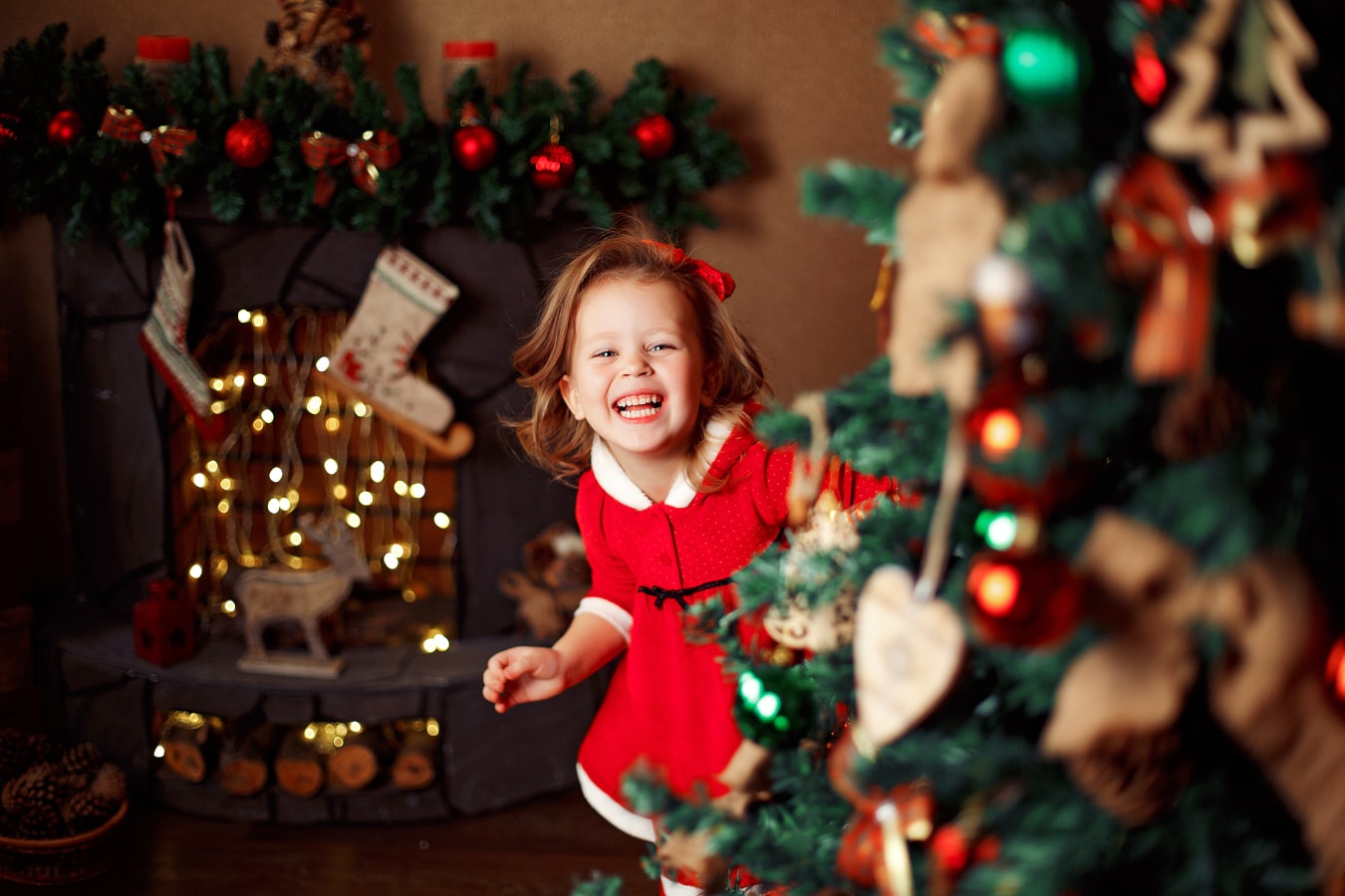 Une petite fille joyeuse dans une robe rouge regarde de derrière le sapin de Noël, en riant, elle est heureuse.